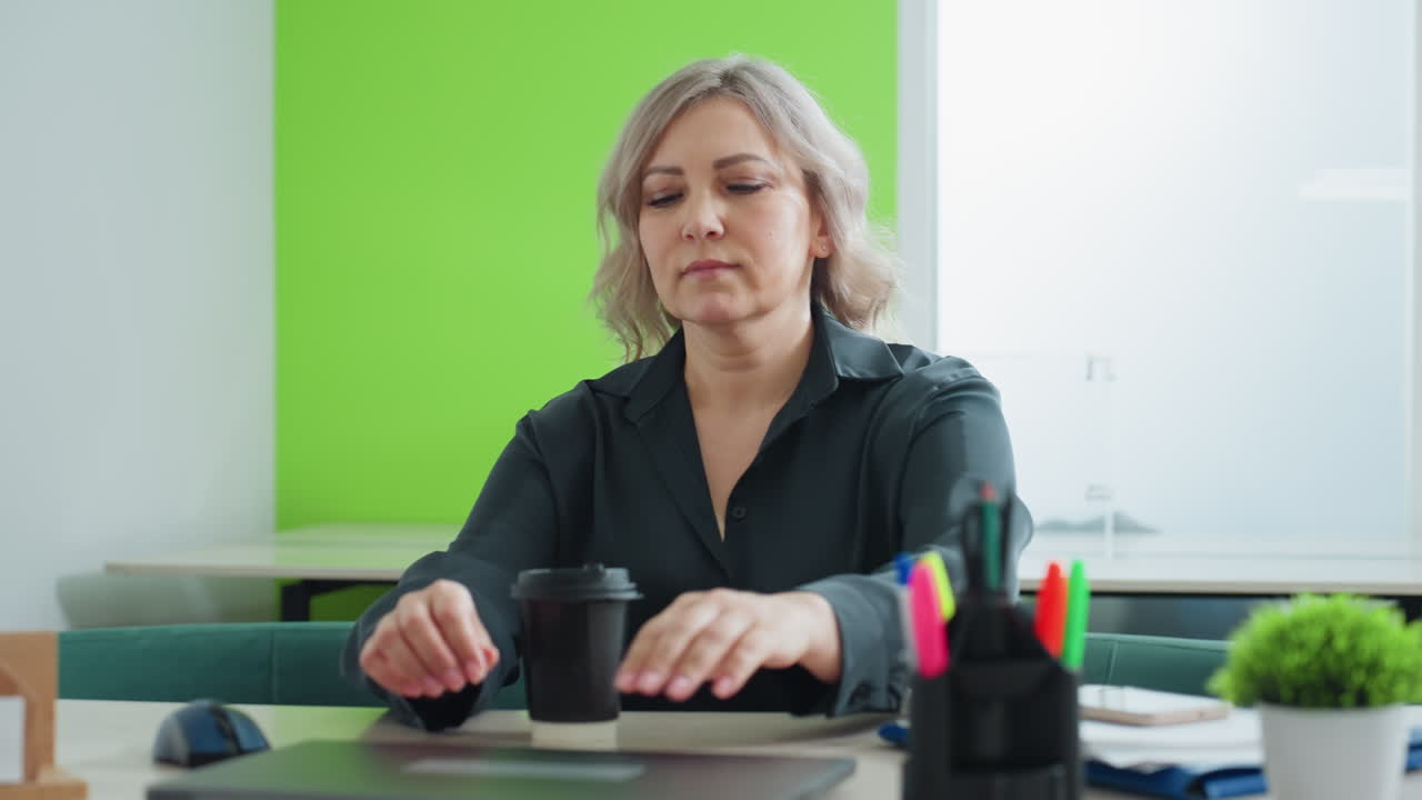 Professional real estate agent in dark blouse holds folder with documents, looking down thoughtfully while preparing to start work in bright office with green accent wall and modern furnishings