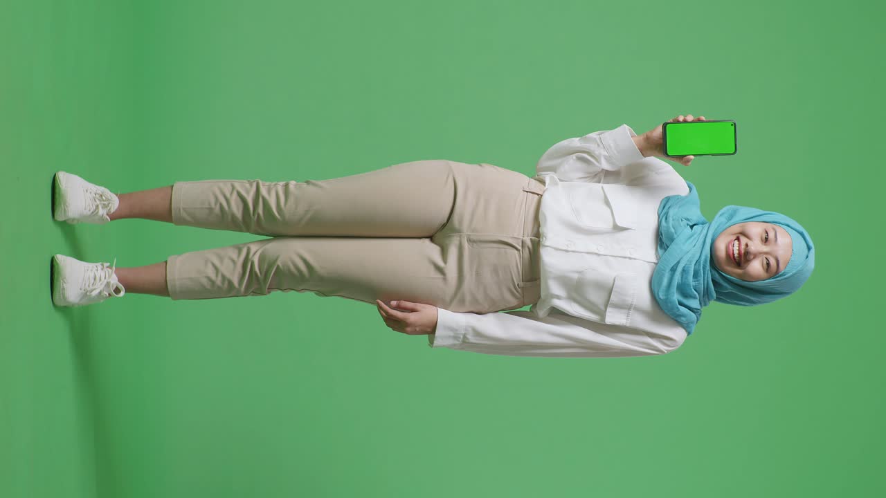 아시아 무슬림 여성이 미소 짓고 녹색 스크린 스마트폰을 카메라에 보여주면서 녹색 화면 배경 스튜디오에 서 있습니다.