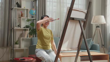 artista asiática con un pincel limpia el sudor antes de cruzar los brazos y sonriendo después de terminar de pintar en el lienzo