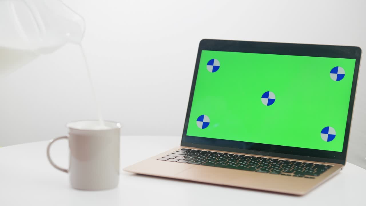 primer plano vertiendo leche en una taza en cámara lenta con una computadora portátil chromakey de pie en la mesa. mujer joven caucásica irreconocible preparando una bebida saludable navegando por internet en un dispositivo de pantalla verde