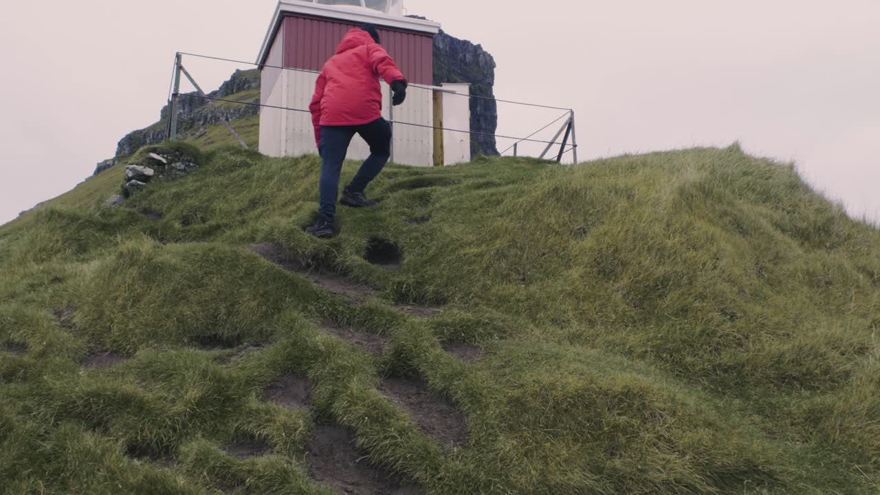 hombre con chaqueta roja sube una montaña verde y llega al faro, kalsoy en la cima de las islas feroe en un día nublado