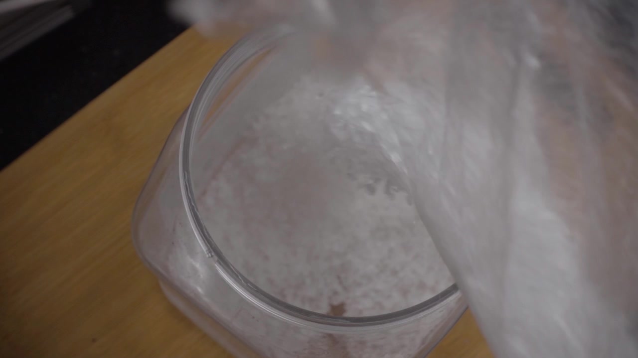 Close-up macro de sal granulado sendo despejado em recipiente de plástico sobre mesa de madeira. Imagens de ingrediente alimentar destacando finos cristais de sal, armazenamento de cozinha e foco seletivo.