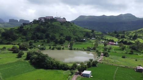 Aerial-view-of-Nil-Parvat-sacred-temple-on-the-edges-of-Brahmagiri-hills-with-the-view-of-Trimbakeshwar-town-and-Sahyadri-mountains-under-an-overcast-sky-in-monsoon,-Nashik,-Maharashtra,-India