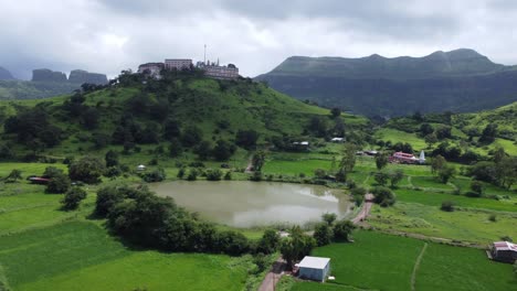 Vista-Aérea-Del-Templo-Sagrado-Nil-Parvat-En-Los-Bordes-De-Las-Colinas-Brahmagiri-Con-La-Vista-De-La-Ciudad-De-Trimbakeshwar-Y-Las-Montañas-Sahyadri-Bajo-Un-Cielo-Nublado-En-Monzón,-Nashik,-Maharashtra,-India