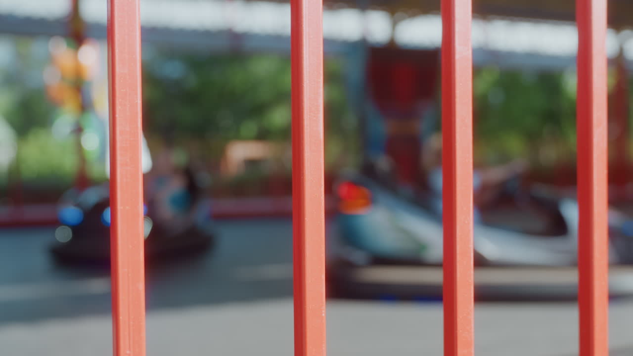 Vista distante di un'attrazione di autoscontri recintata nel parco nel weekend attraverso sbarre verticali rosse, autoscontri sfocate che sfrecciano sulla pista, tettoie colorate e vegetazione, prospettiva spontanea dello spettatore, genitore e figlio.