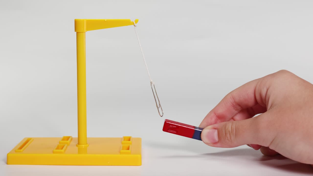 A hand manipulates a magnet near a suspended paperclip, illustrating magnetic forces in a controlled environment with clear lighting