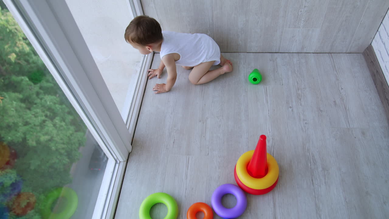 Baby boy plays with pyramid on the floor at panoramic window. Kid notices a car riding on the street and leans to the window. Top view.