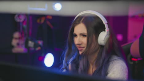 Tense female gamer wearing white headphones focused on screen while reacting with visible displeasure after losing game, seated in colorful studio with condenser microphone in front