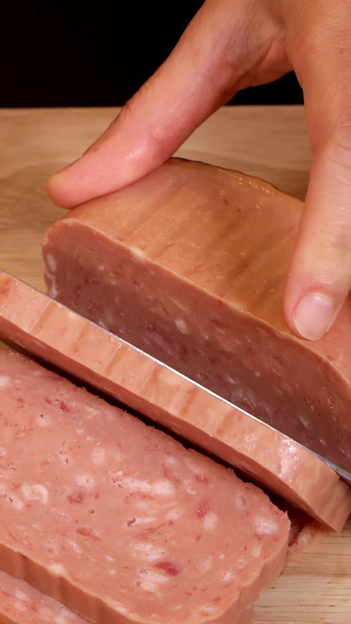 Una mano usa un coltello da cucina per affettare un blocco di salume lavorato in pezzi uniformi su un tagliere di legno sotto una luce soffusa.