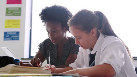 Weibliche-Gymnasiallehrerin-Mit-Digitalem-Tablet,-Die-Mädchen-In-Uniform-Eins-Zu-Eins-Unterricht-Am-Schreibtisch-Gibt