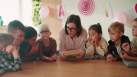 una chica rubia con un peinado de bob con gafas en una camisa blanca lee un libro a los niños en edad preescolar mientras ella decide en el suelo sobre almohadas especiales con los niños que la escuchan