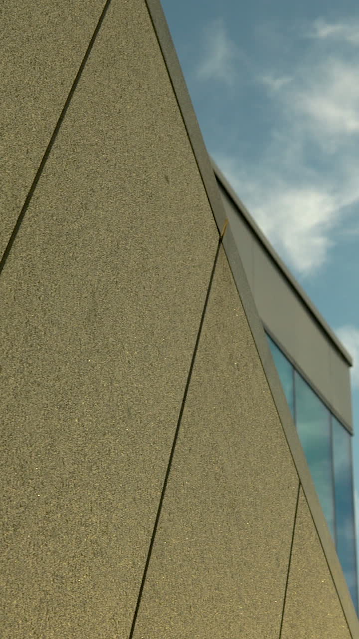 Uma fotografia vertical mostrando os detalhes geométricos de uma fachada de edifício contemporâneo, com painéis de betão texturizados e janelas de vidro sob um céu claro.