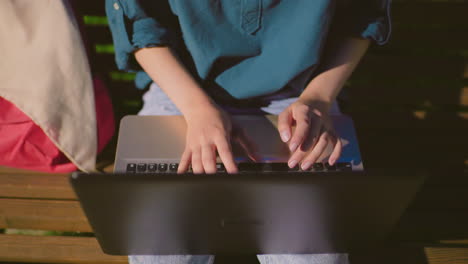 close-up of woman using laptop outdoors at night, fingers typing on keyboard with white reflection on screen, background features soft blur