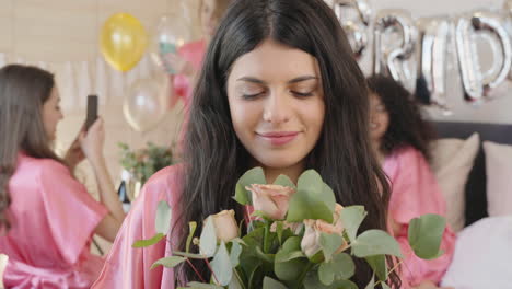 brunette woman holding and smelling a bouquet, wearing pink silk nightdress, smiling and looking at camera 2