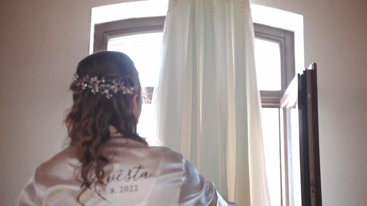 novia en bata admirando su vestido de novia colgando de la viga del techo