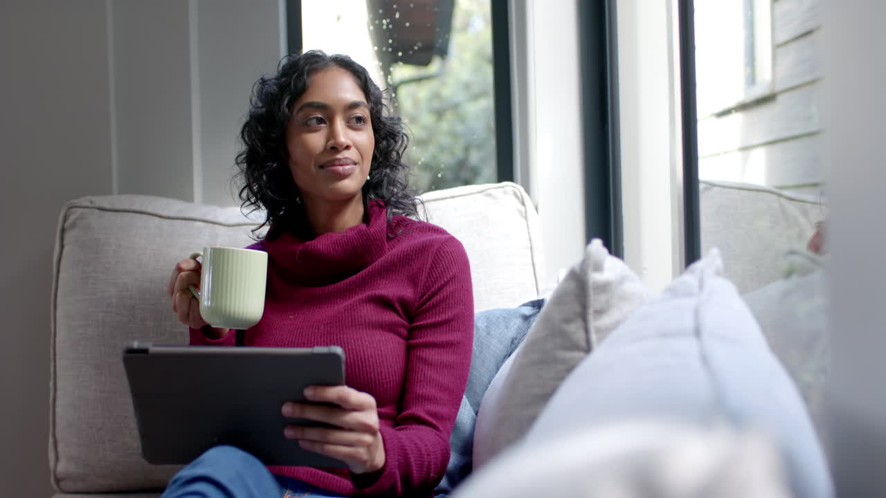 Biracial woman sitting on sofa using tablet and drinking coffee at home, slow motion