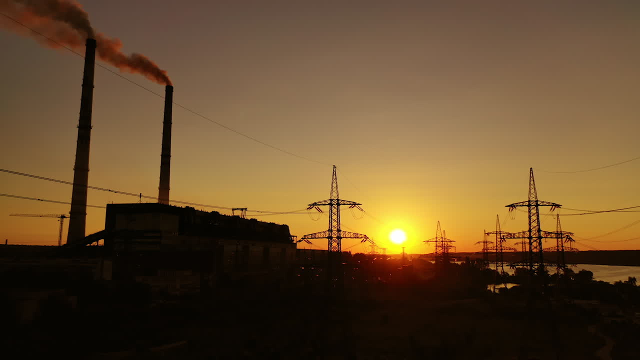 Power station at sunset. Transmission lines near industrial factory with pipes. High voltage electric lines at dusk. Dark smoke from industrial pipes in the evening.
