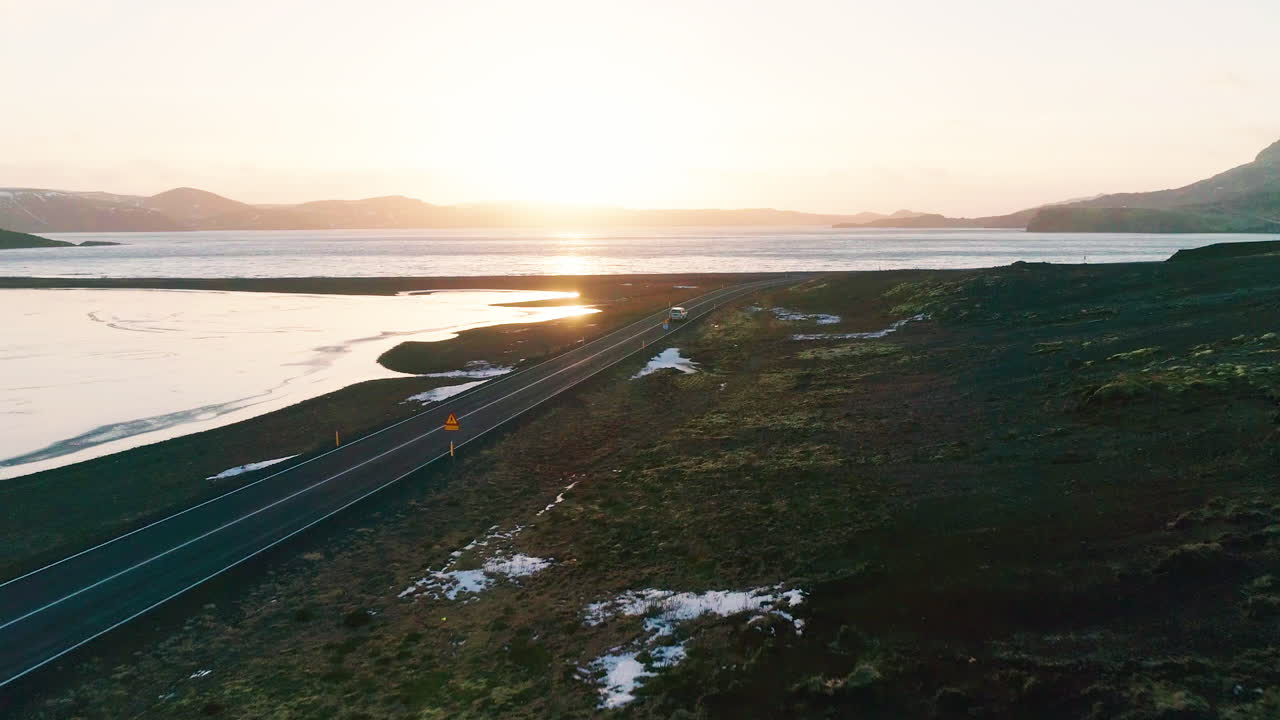 vehículo que conduce el lago kleifarvatn largo camino curvo rodeado de montañas de lava al atardecer, vista aérea descendente hacia adelante