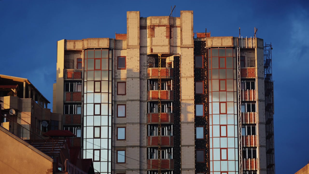 Immeuble résidentiel de moyenne hauteur en construction, avec des sections de briques apparentes, une façade en béton non finie, des panneaux vitrés installés et des échafaudages visibles sur le côté droit