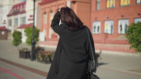 lady in black coat and white sweater underneath adjusts her hair back while carrying black handbag in sunny urban setting with red brick building and green trees in the background