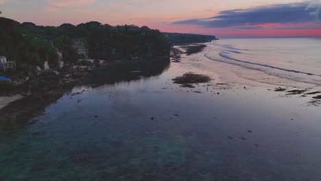 Drohnenaufnahme-Eines-Wunderschönen-Rosafarbenen-Sonnenuntergangs-Am-Strand-Von-Bingin-Auf-Bali,-Mit-Spiegelungen-Des-Sonnenuntergangs-Und-Kalksteinfelsen-Im-Ruhigen-Wasser