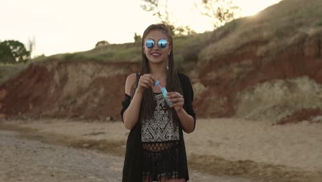 joven hipster con rastas alegremente haciendo pompas de jabón en la playa por la noche