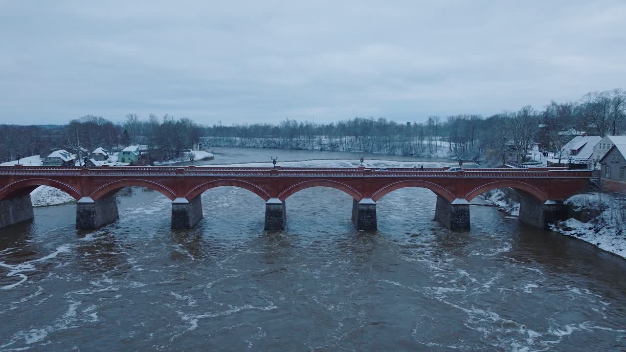 vista aérea de los rápidos del río venta durante las inundaciones de invierno, viejo puente de ladrillo rojo, kuldiga, letonia, día de invierno nublado, amplia toma de drone avanzando