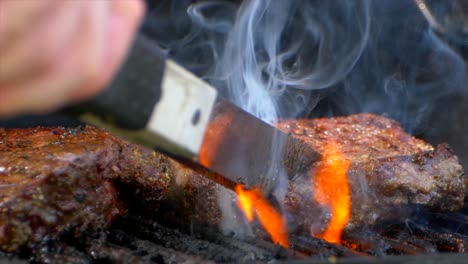 a pair of meat tongs slide a nearly cooked juicy rib eye steak on a grill and presses on it as flames shoot up in slow motion