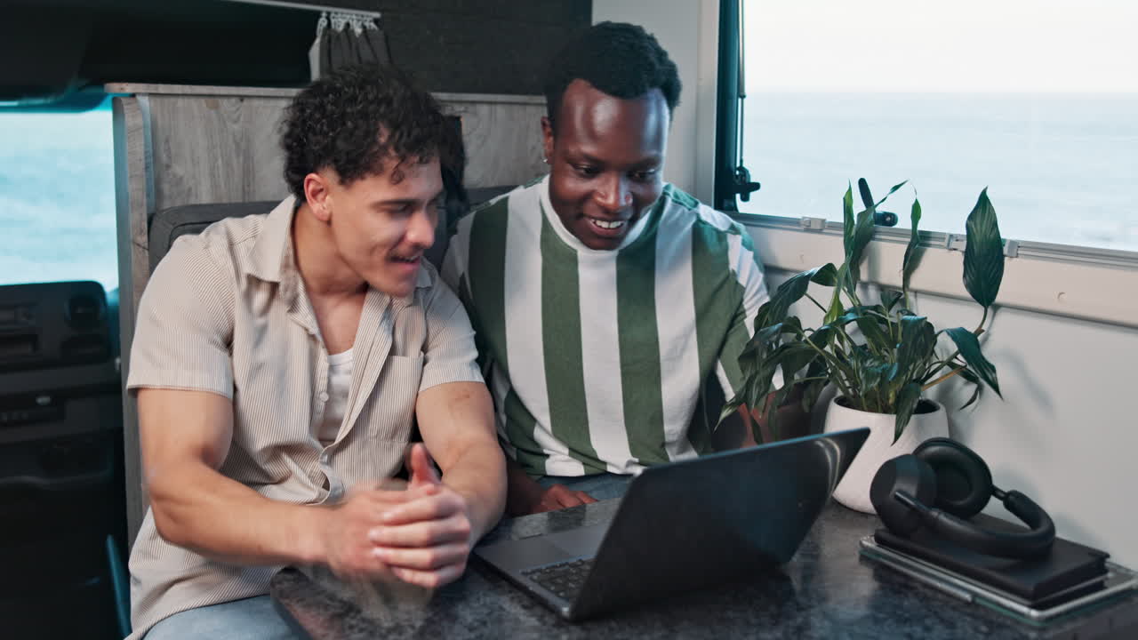 Two Young Men Smiling and Talking While Using a Laptop in a Camper Van