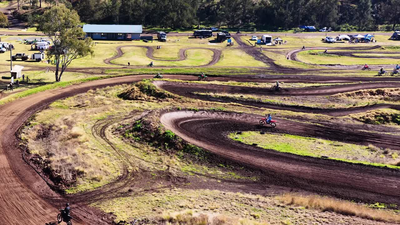 Motocross rider speeds along winding dirt track in bright daylight, wide static shot, rural setting