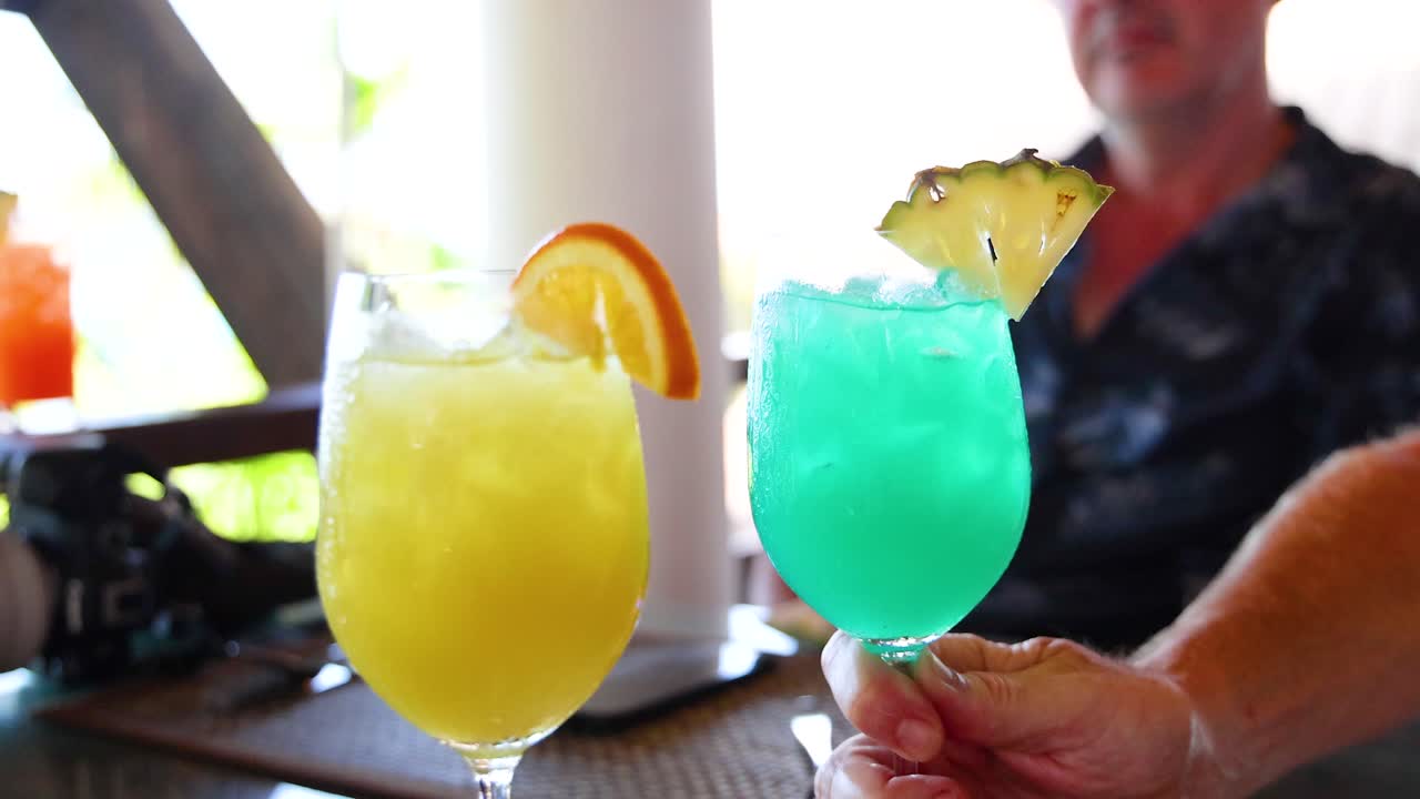 Two vibrant cocktails clinked together in a bright, tropical environment. Sunlight enhances the colorful drinks and relaxed atmosphere