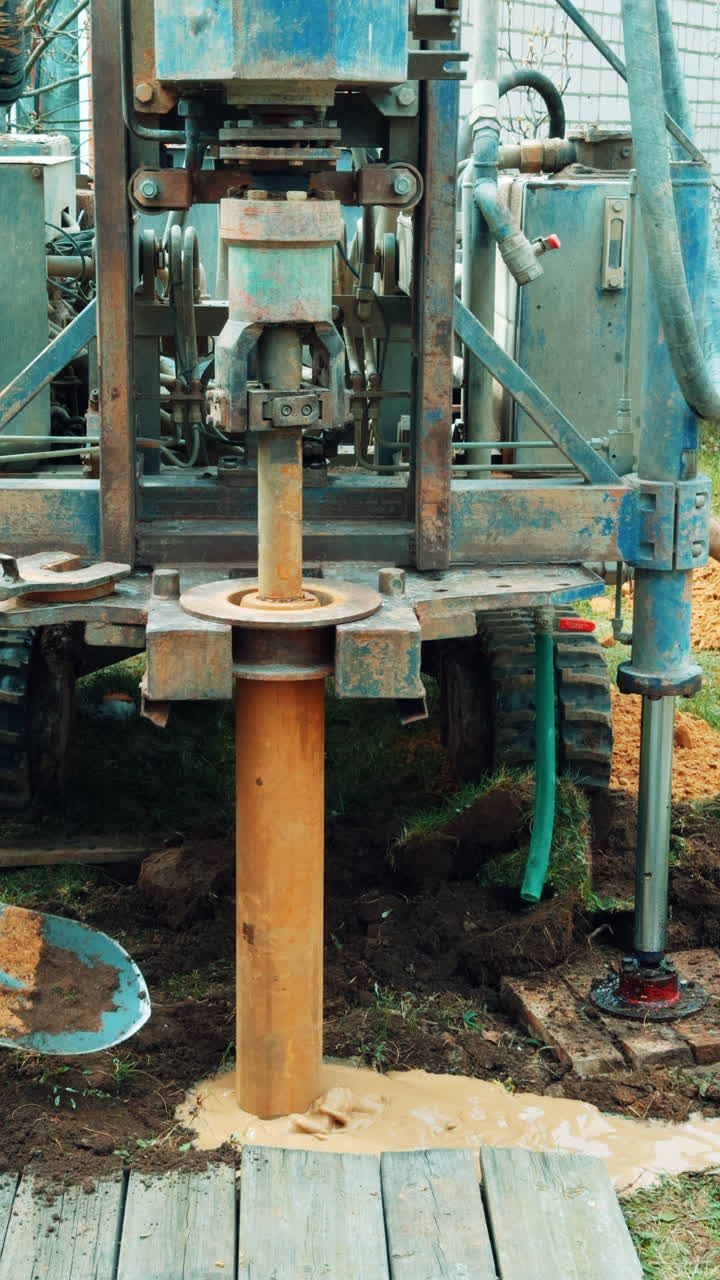 Nozzle of drilling machine rotates in the ground making a well. Worker uses a shovel to move the ground. Vertical video.