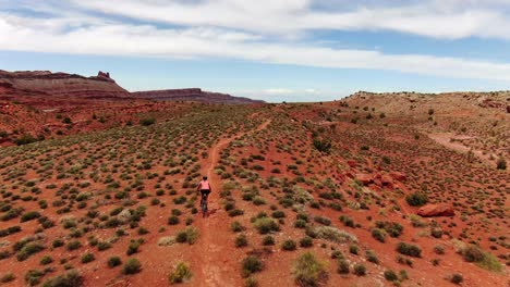 Toma-Aérea-Detrás-De-Una-Mujer-En-Bicicleta-De-Montaña-En-Un-Sendero-Plano-Con-Arenisca-Roja-Del-Desierto