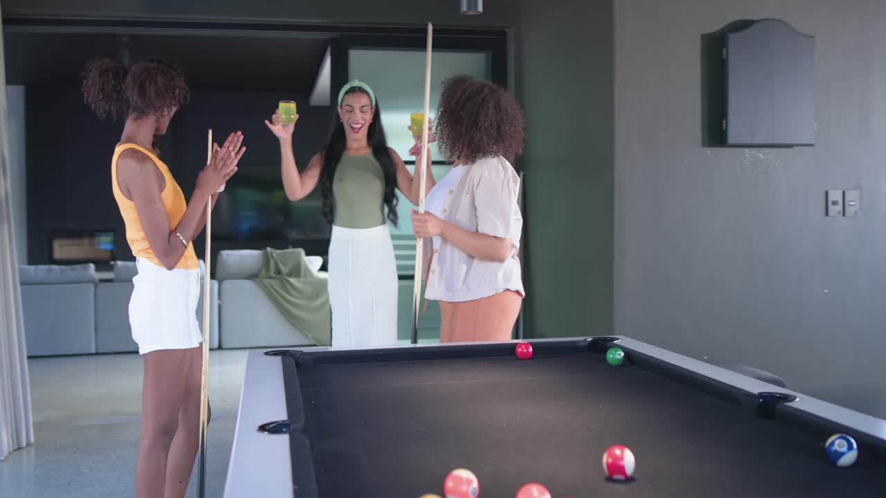 Donne afroamericane che giocano a biliardo a casa mentre salutano un'amica che entra con bevande per socializzare