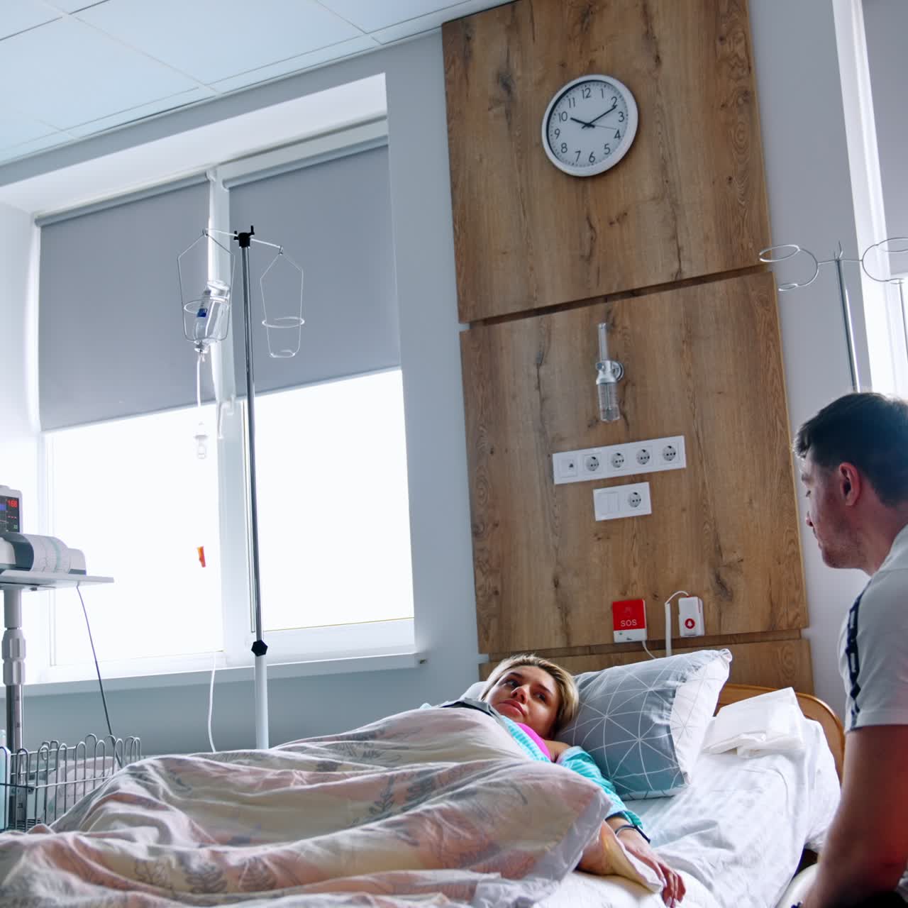 Pregnant woman lies in the hospital bed covered with duvet. Husband sits beside his wife talking and smiling to her. Low angle view