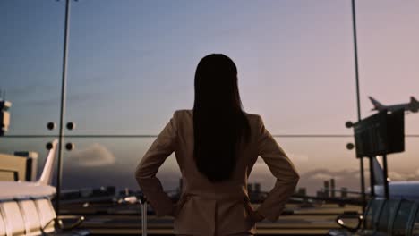 back view of asian businesswoman with rolling suitcase in boarding lounge at the airport, businesswoman with arms akimbo looking around waits for flight, airplane takes off outside the window