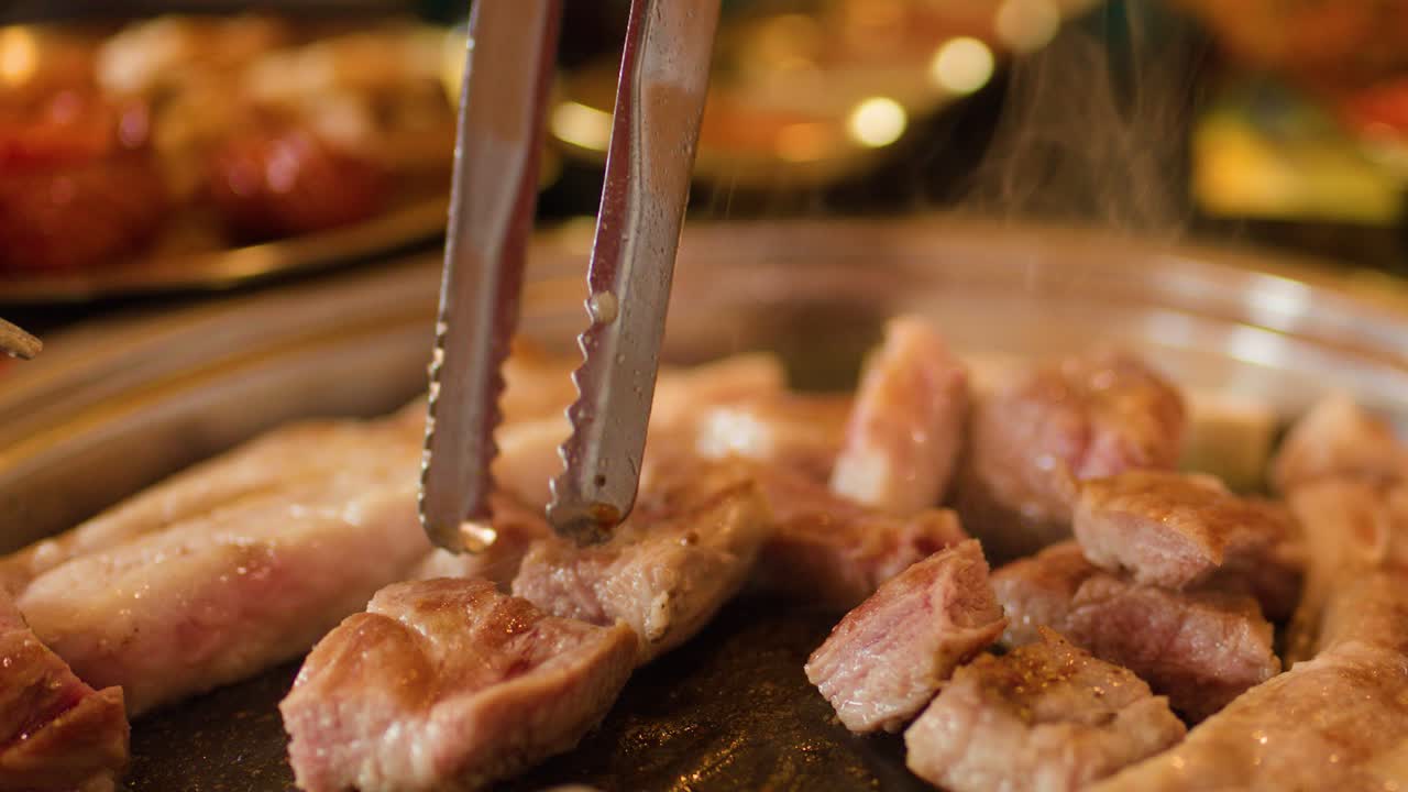 Tongs turn sizzling pork belly on a hot grill, steam rising in warm restaurant light