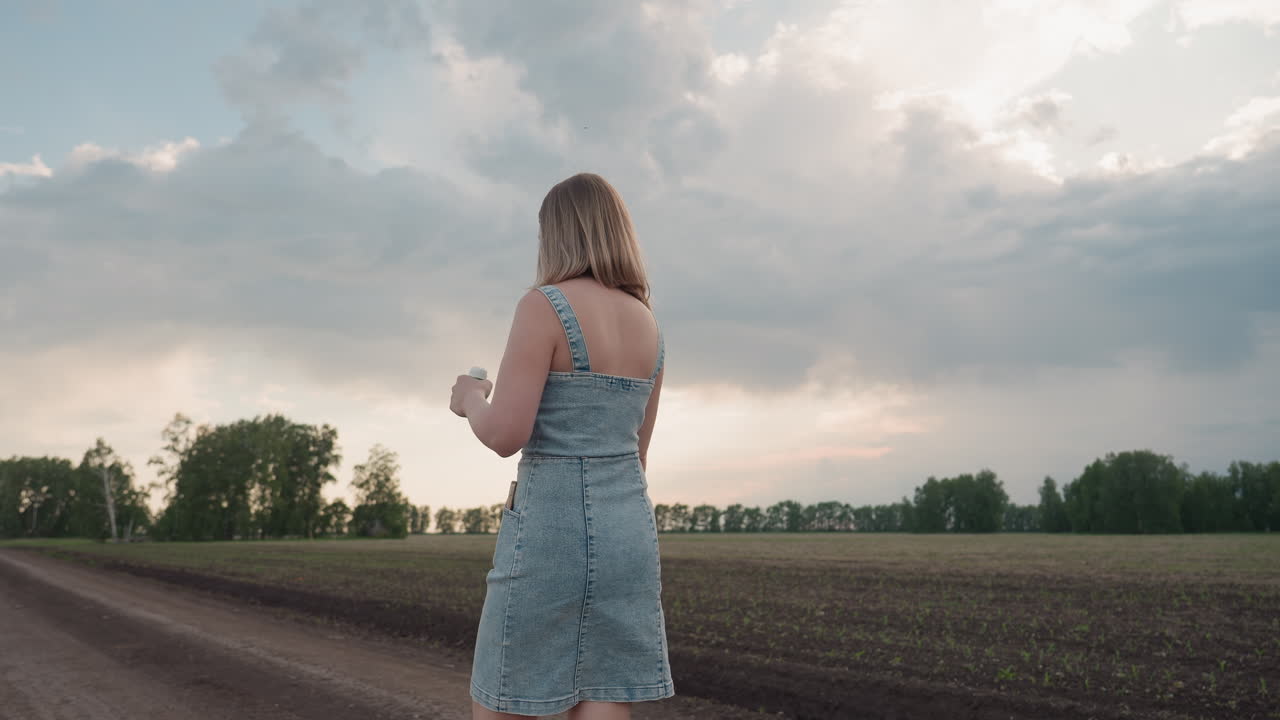 rear view woman strolling across wide farmland holding bubble container smiling under vast cloudy sky during golden hour summer scene with soft sunset glow illuminating profile