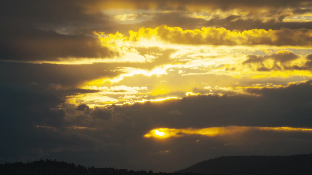 Golden evening sunlight filters through heavy layered storm clouds above a dark distant hillside.