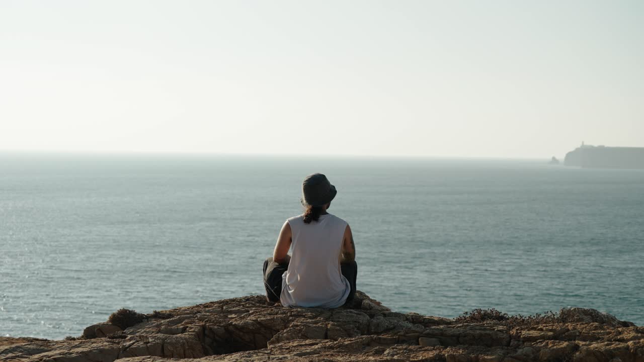 A man sits in stillness atop a rocky cliff, facing the calm sea and distant horizon in Algarve, Portugal