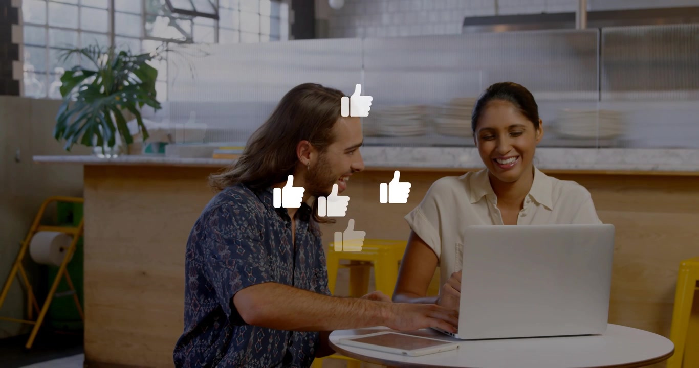 Mand taster på bærbar computer, og kvinde følger med på café, mens de skaber marketinggrafik med tommelfinger op, der mangedobles.