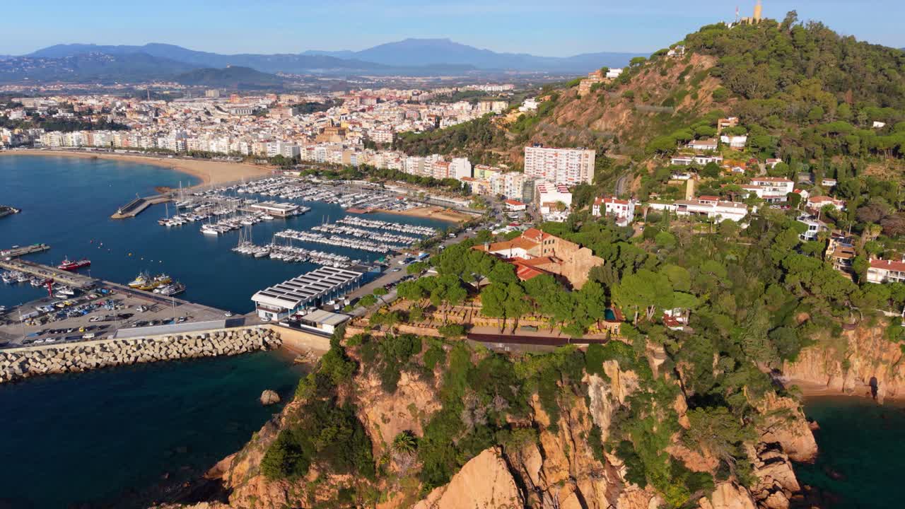 Picturesque aerial view from a drone flying over the coastal town of blanes, revealing the bustling port, beautiful beach, and historic castell de sant joan on a sunny day in costa brava, spain