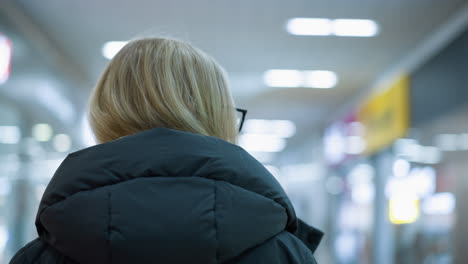 head view of young lady in glasses and black hoodie walking in well-lit mall, she looks around with soft smile, blurred background with other shoppers and glowing lights creating dreamy bokeh effect