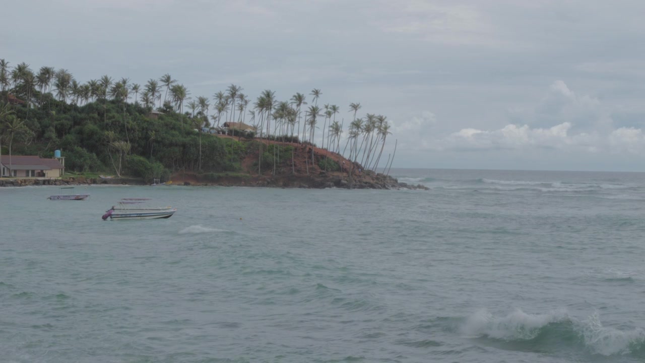 Ampia vista costiera di Mirissa, Sri Lanka, che mostra onde oceaniche agitate, piccole imbarcazioni e un promontorio roccioso coperto di palme sotto un cielo tropicale nuvoloso.