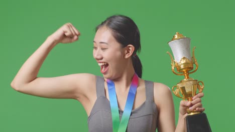 close up of asian woman with a gold medal and trophy flexing her bicep and smiling to camera on green screen background in the studio