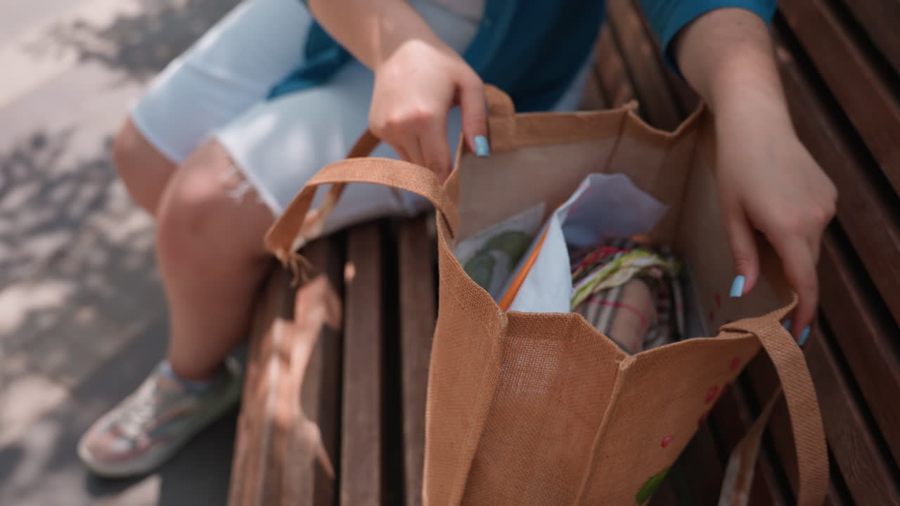Close up of girl on wooden bench placing embroidery materials and fabric into brown tote bag under sunlight, preparing to leave after creative activity, calm outdoor atmosphere