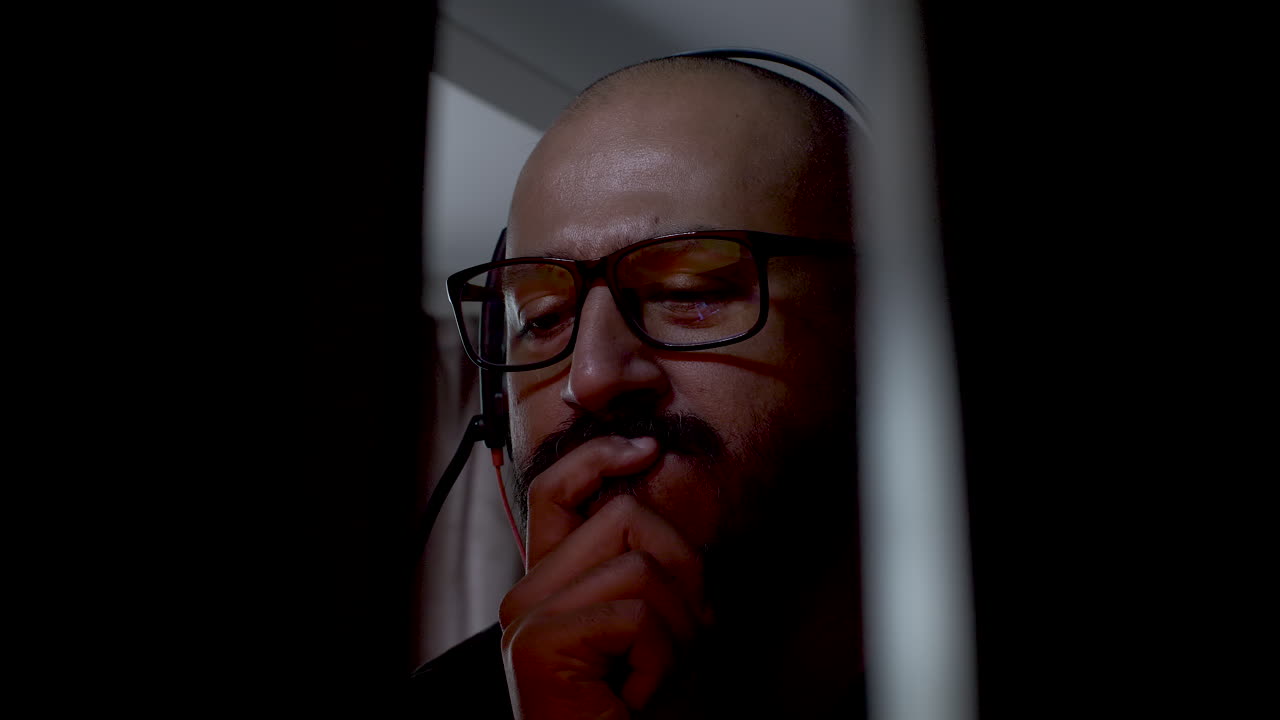 hombre calvo con gafas, bigote y auriculares pensando y mirando una pantalla de computadora que muestra colores brillantes mientras se lleva la mano a la boca