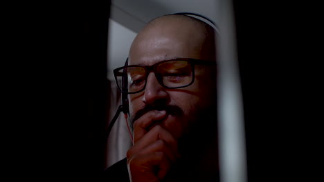 bald man with glasses, moustache and headset thinking and looking at a computer screen that flashes bright colors while he holds his hand in his mouth