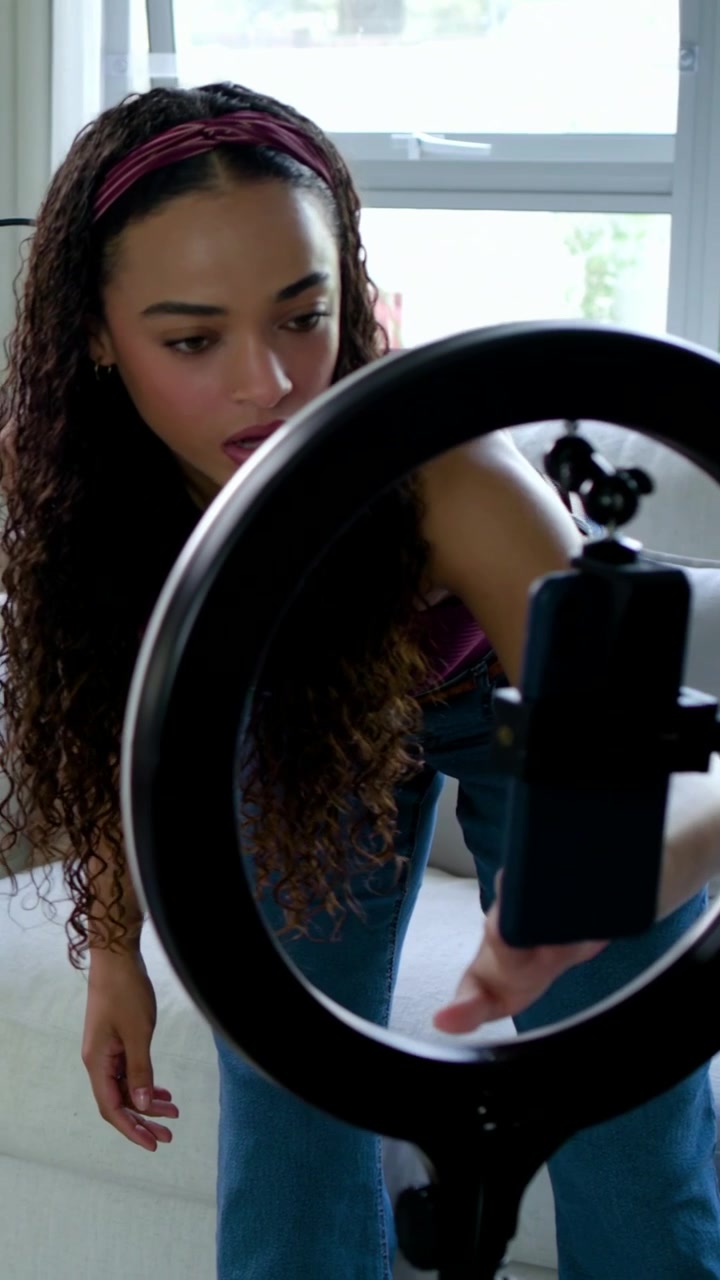Vertical video: Entering woman creator in maroon adjusting ring light, phone, mic at home to record