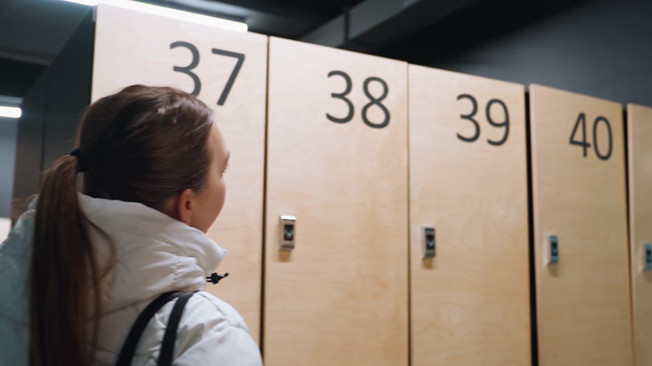 Player arrives at badminton club lockers wearing white puffer coat and carrying large sports bag, scans numbered wooden lockers, pauses with thermos, selects locker and prepares to unlock, calm
